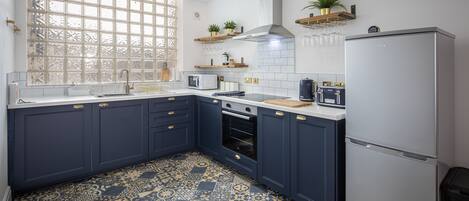 Une cuisine avec des meubles de couleur bleu foncé, un réfrigérateur blanc, un carrelage au motif décoratif et une fenêtre ornée d’une grille.