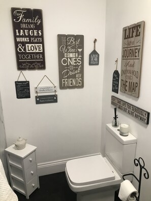 A bathroom with a toilet, a small cabinet, and a wall-mounted shelf with a candle and a tissue roll.