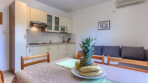 Ett kompakt kök med vita skåp, en mikrovågsugn och en spis. Det finns ett matbord med en ananas och bananer på, och en grå soffa i bakgrunden.