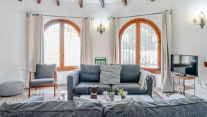 Ein gemütliches Wohnzimmer mit einer Couch, einem Sessel, einem Hocker, einem Couchtisch mit Pflanzen und einem Fernseher auf einem Lowboard.