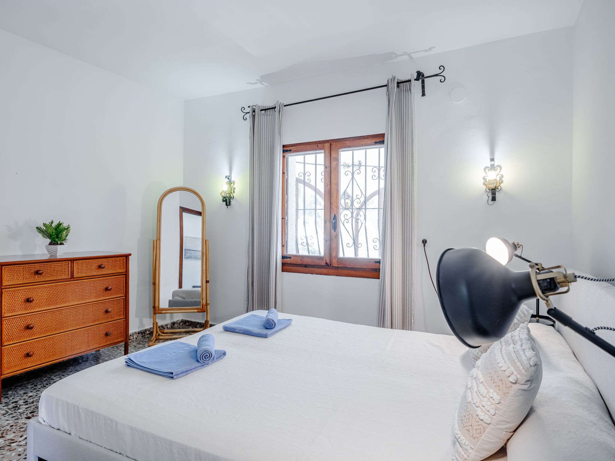 A bedroom with a bed, a dresser, a mirror, a window with curtains, and wall sconces.