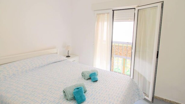 A bedroom with a bed, two towels, a nightstand, a lamp, and a sliding door to a balcony. - Rainbow by Interhome (Benidorm)