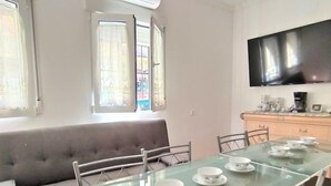 A dining area with a glass table set for four, a grey sofa, and a TV mounted on the wall. - Rainbow by Interhome (Benidorm)