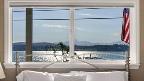 A bed with white linens and a view of a lake with mountains in the background through a window with a metal frame and a flag. - Waterviews, Gameroom, close to DT, & Ferries! (Port Orchard)