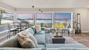 A modern living room with a large window overlooking a scenic view, a gray sofa, a dining area with chairs, and a ladder against the wall. - Waterviews, Gameroom, close to DT, & Ferries! (Port Orchard)