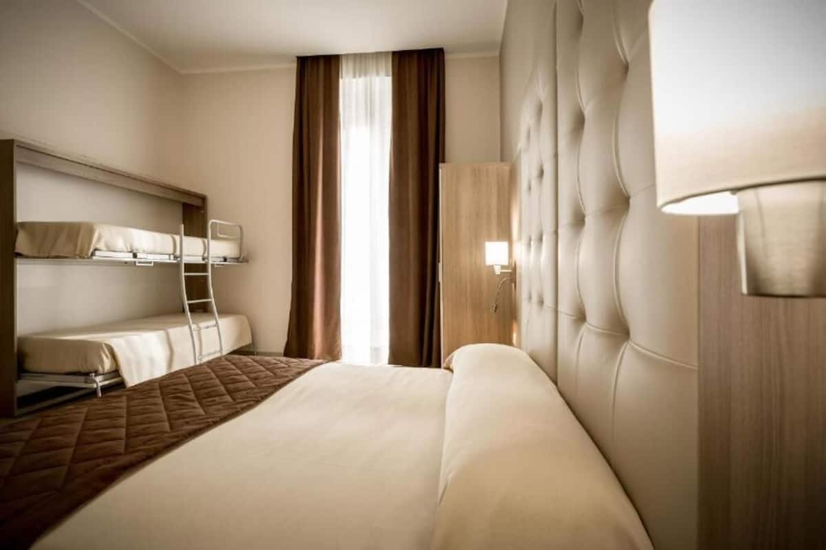 A hotel room with a large bed, a bunk bed, a wardrobe, and a window with curtains.