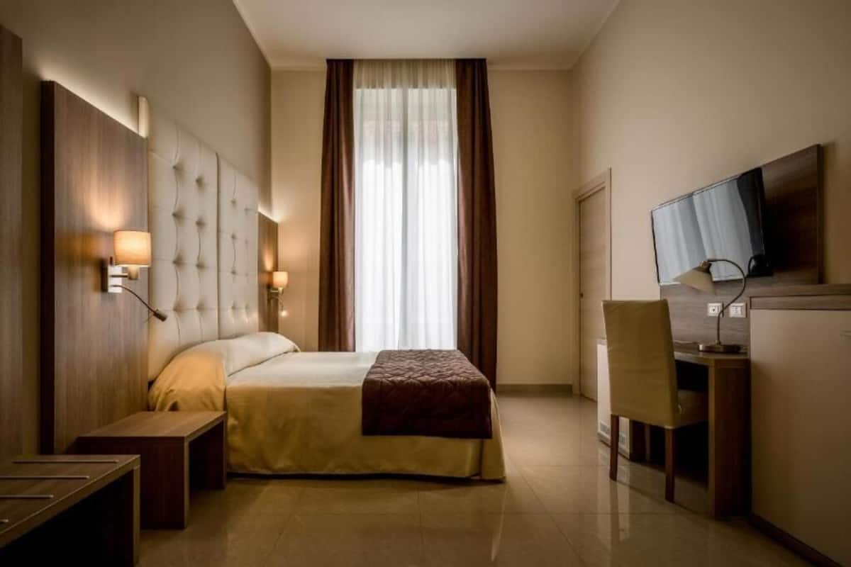 A hotel room with a large bed, a desk with a chair, a television, and a window with curtains.