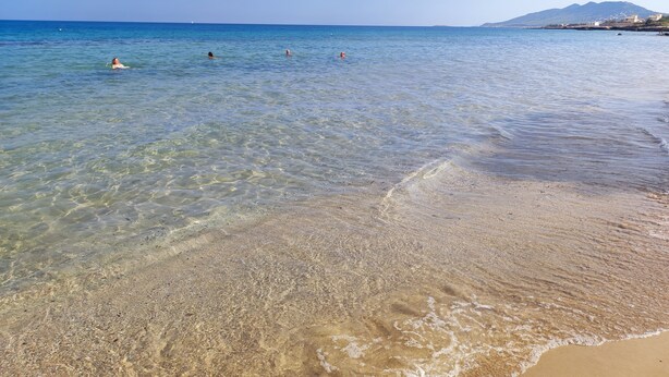 Una spiaggia con acque limpide e poco profonde, un litorale sabbioso e una montagna in lontananza.