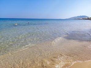 Een strand met helder ondiep water, een zandstrand en een berg in de verte.