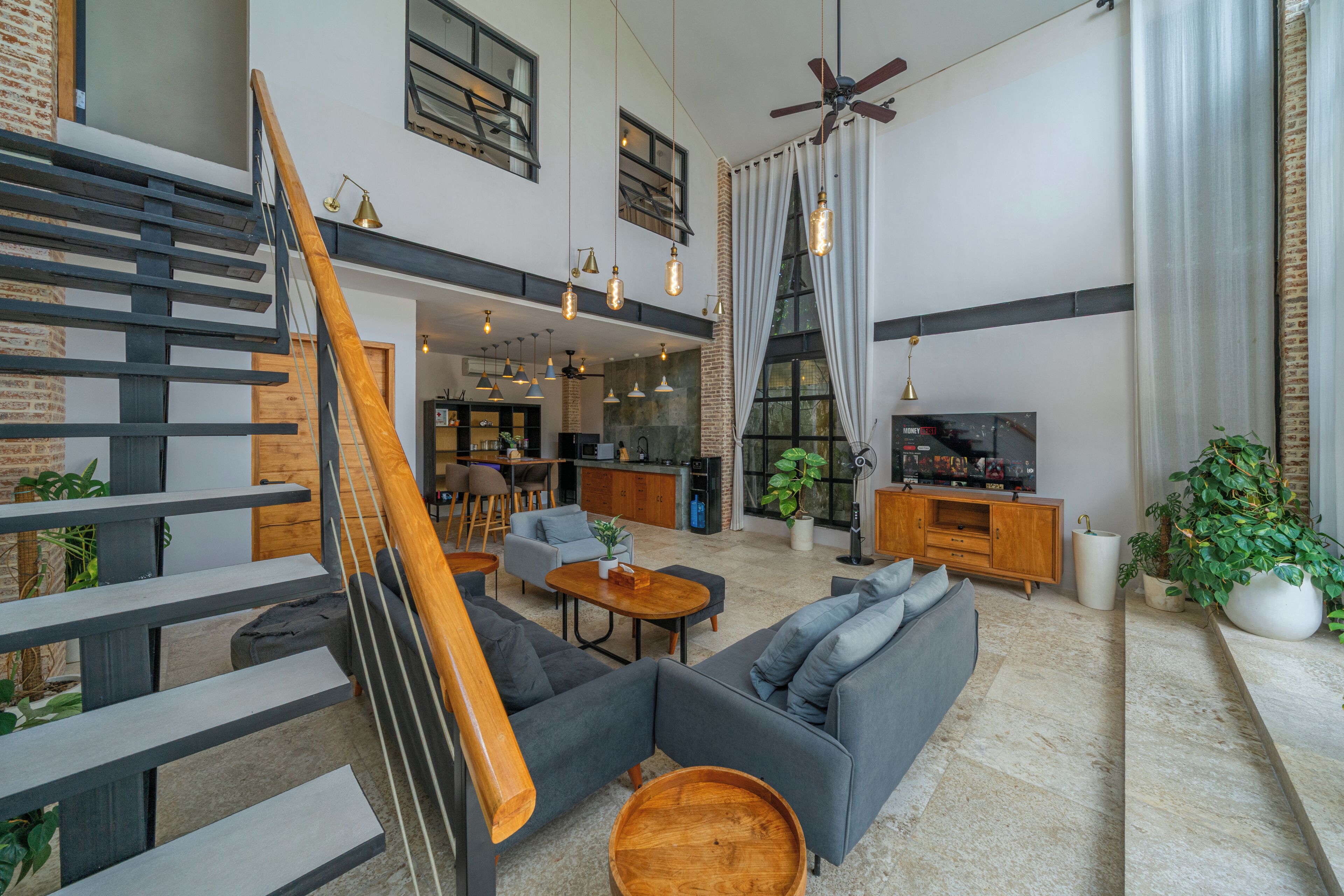 A spacious living area with a staircase, seating, a television, and a kitchen in the background.