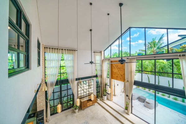 A spacious room with large windows, a ceiling fan, and a view of a swimming pool. - Villa Upeksha (Canggu)