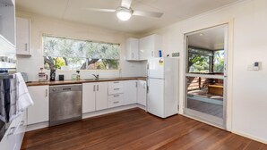 Una cocina con armarios blancos, lavavajillas y refrigerador. Hay una ventana con vistas a árboles y una puerta que da a un patio.
