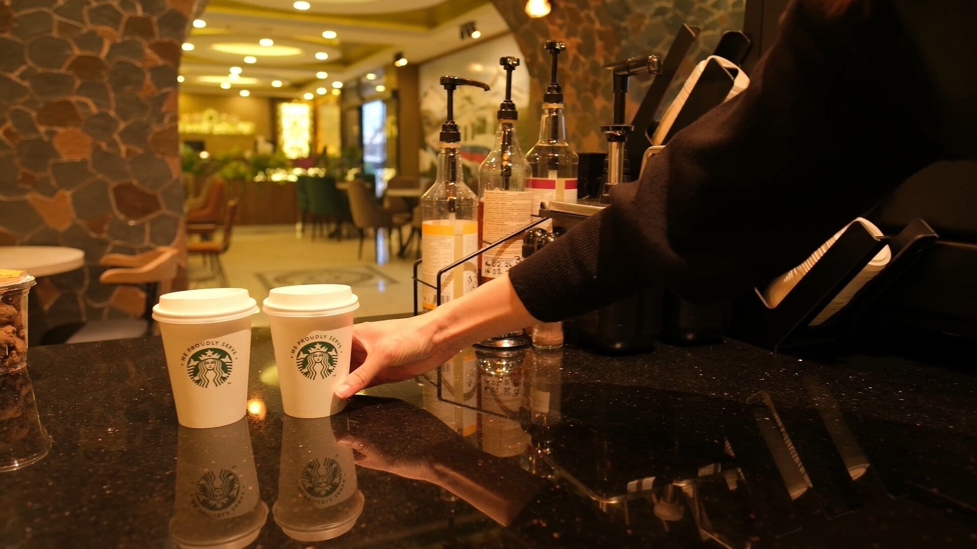 Un barista colocando tazas de Starbucks sobre un mostrador en un ambiente interior bien iluminado con paredes de piedra y una decoración moderna.
