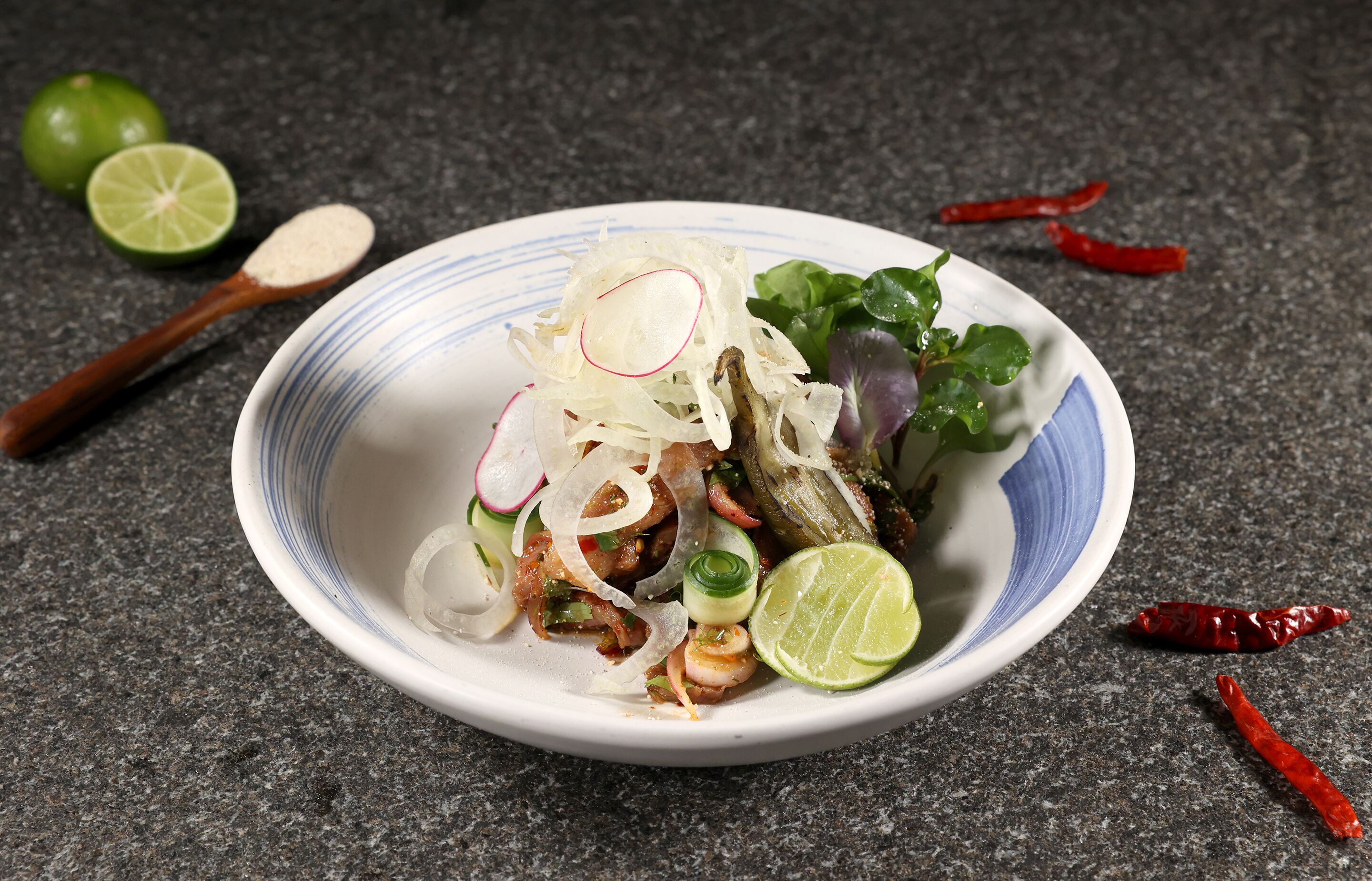 A bowl of salad with lime, radish, and chili peppers on a granite countertop.