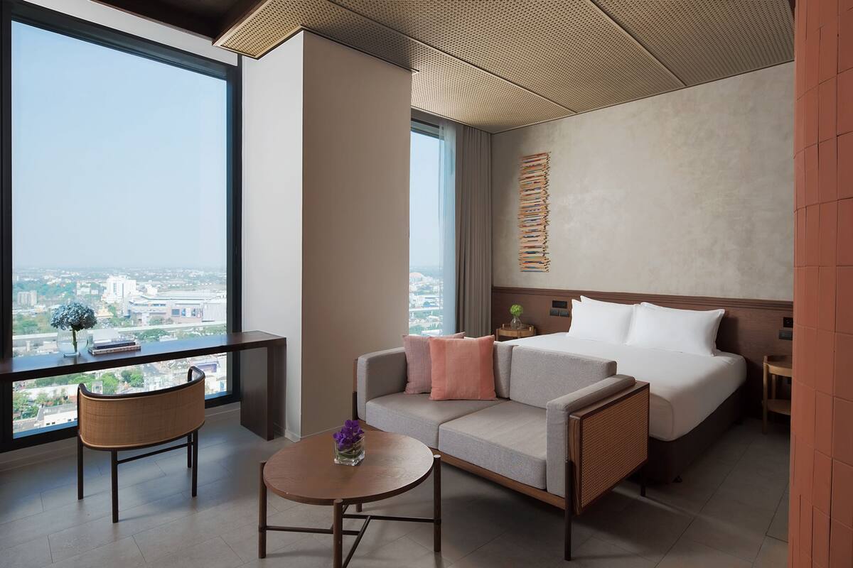 A modern hotel room with a large window, a sofa, a bed, a small table, and a decorative plant.