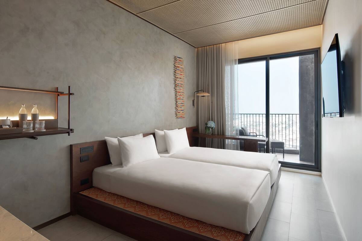 A modern hotel room with two beds, a large window, a flat-screen TV, and a balcony with a view.