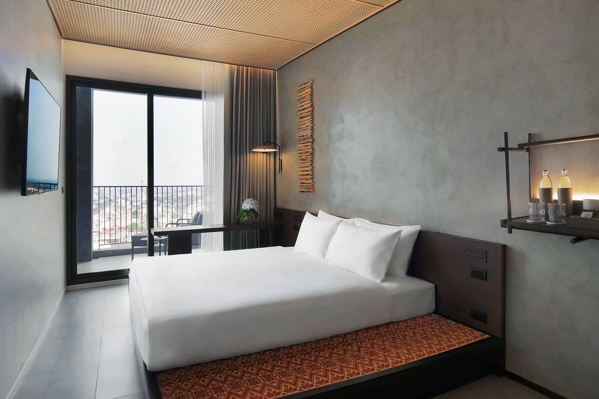 A modern hotel room with a large bed, a TV mounted on the wall, a balcony with a view, and a shelf with bottles.