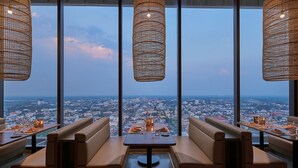 Un restaurante con vistas a la ciudad, que cuenta con mesas altas, asientos acolchados y lámparas de mimbre colgantes.