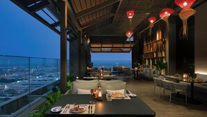 Un área de comedor en la azotea con vista a la ciudad, mesas dispuestas con platos y cubiertos, y faroles rojos colgantes.
