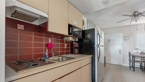Cocina compacta con estufa a gas, microondas y refrigerador, zona de comedor con mesa y sillas, y ventilador de techo.