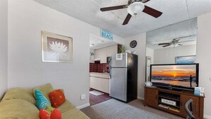 Una sala de estar con sofá, televisor, ventilador de techo y una cocina al fondo.