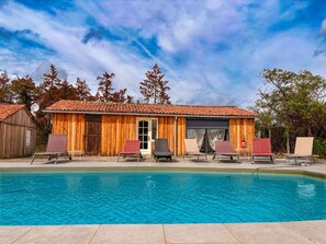 A wooden cabin with a tiled roof, a pool, and lounge chairs.