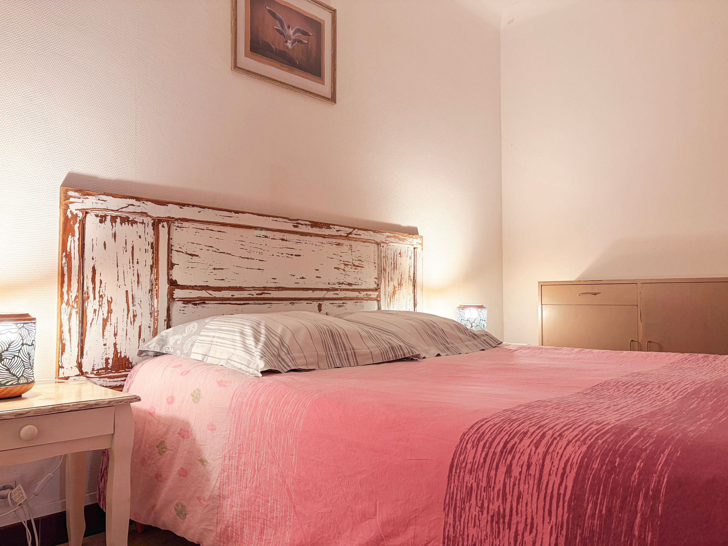 A bedroom with a bed, a bedside table, a nightstand, and a wall-mounted picture.