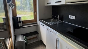 A compact kitchen with white cabinets, a black countertop, a built-in microwave, and a window overlooking a forested area.