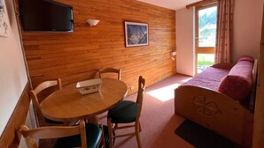 Une pièce chaleureuse avec un mur en bois, une table ronde entourée de chaises, un lit, un téléviseur et une fenêtre avec des rideaux.