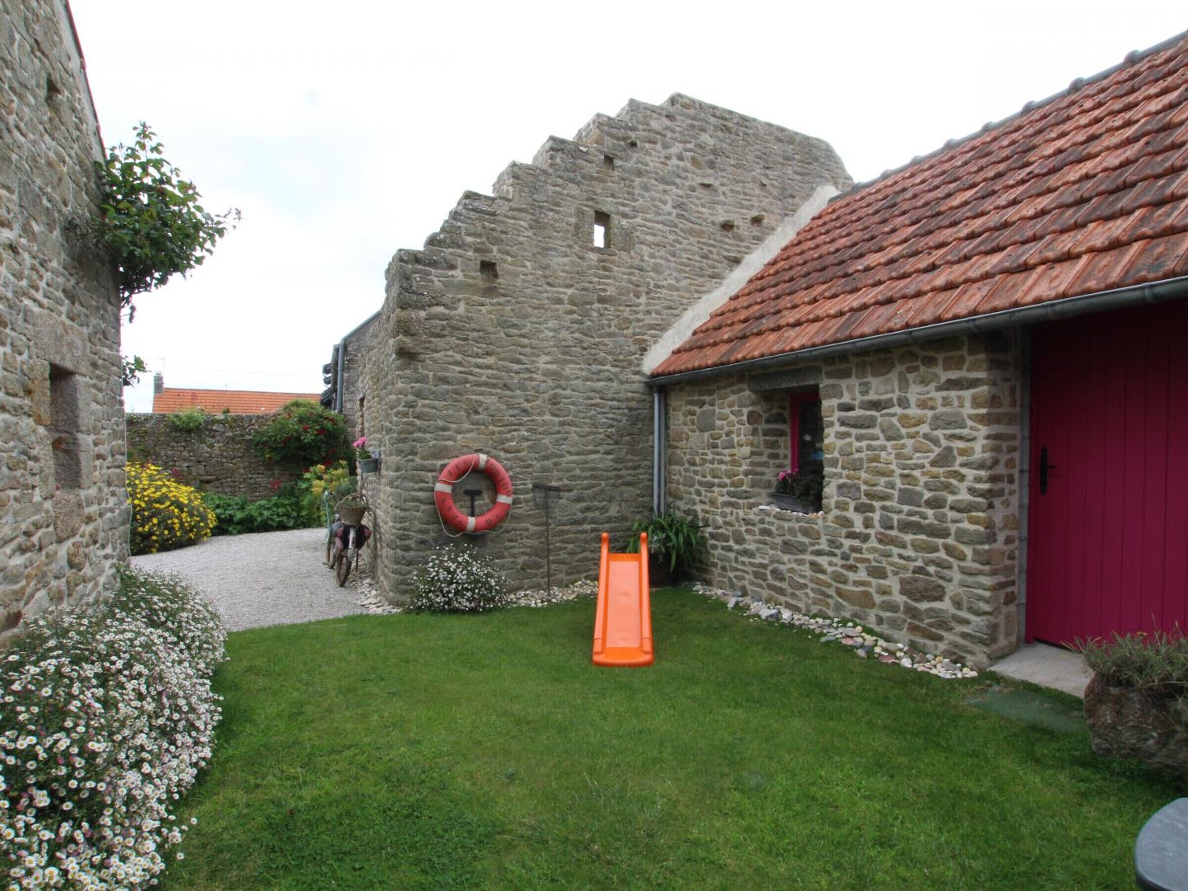 Un bâtiment en pierre avec une porte rouge, une cour verdoyante et une petite structure de jeu orange.
