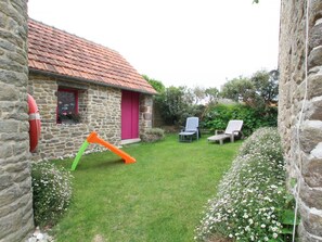 A stone house with a red-tiled roof, a pink door, and a small garden with two lawn chairs and a slide.