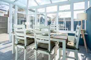 Une véranda avec une table à manger pour quatre personnes, entourée de chaises blanches agrémentées de coussins.