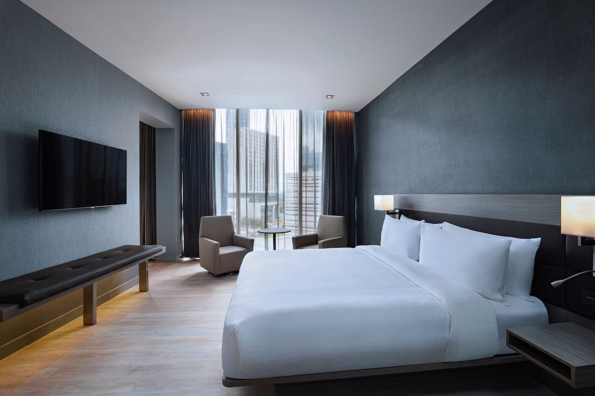 A modern hotel room with a large bed, a flat-screen TV mounted on the wall, a bench, two armchairs, a small table, and a view of the city through large windows.