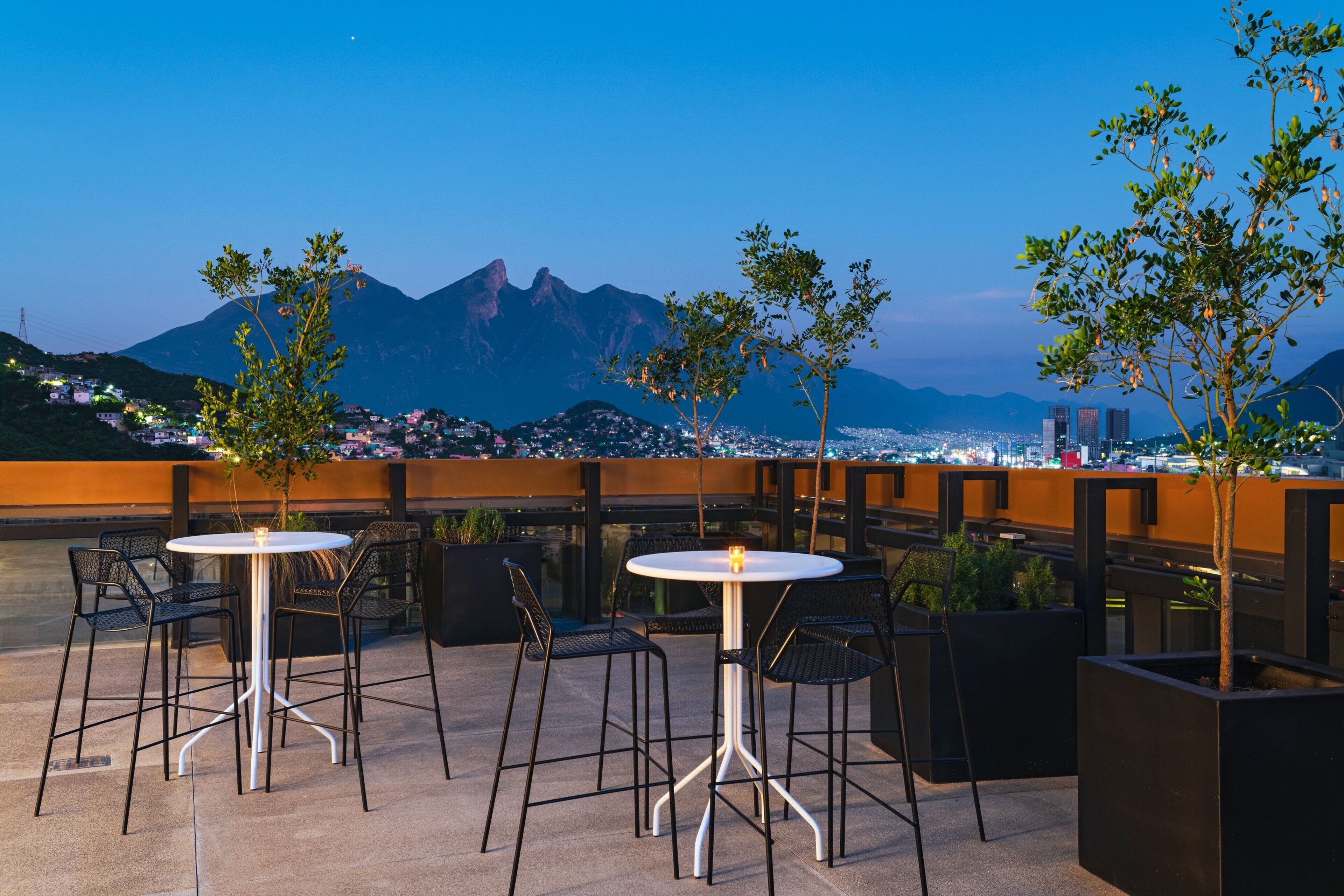 An outdoor terrace with round tables and chairs, surrounded by potted plants, overlooking a cityscape and mountains.