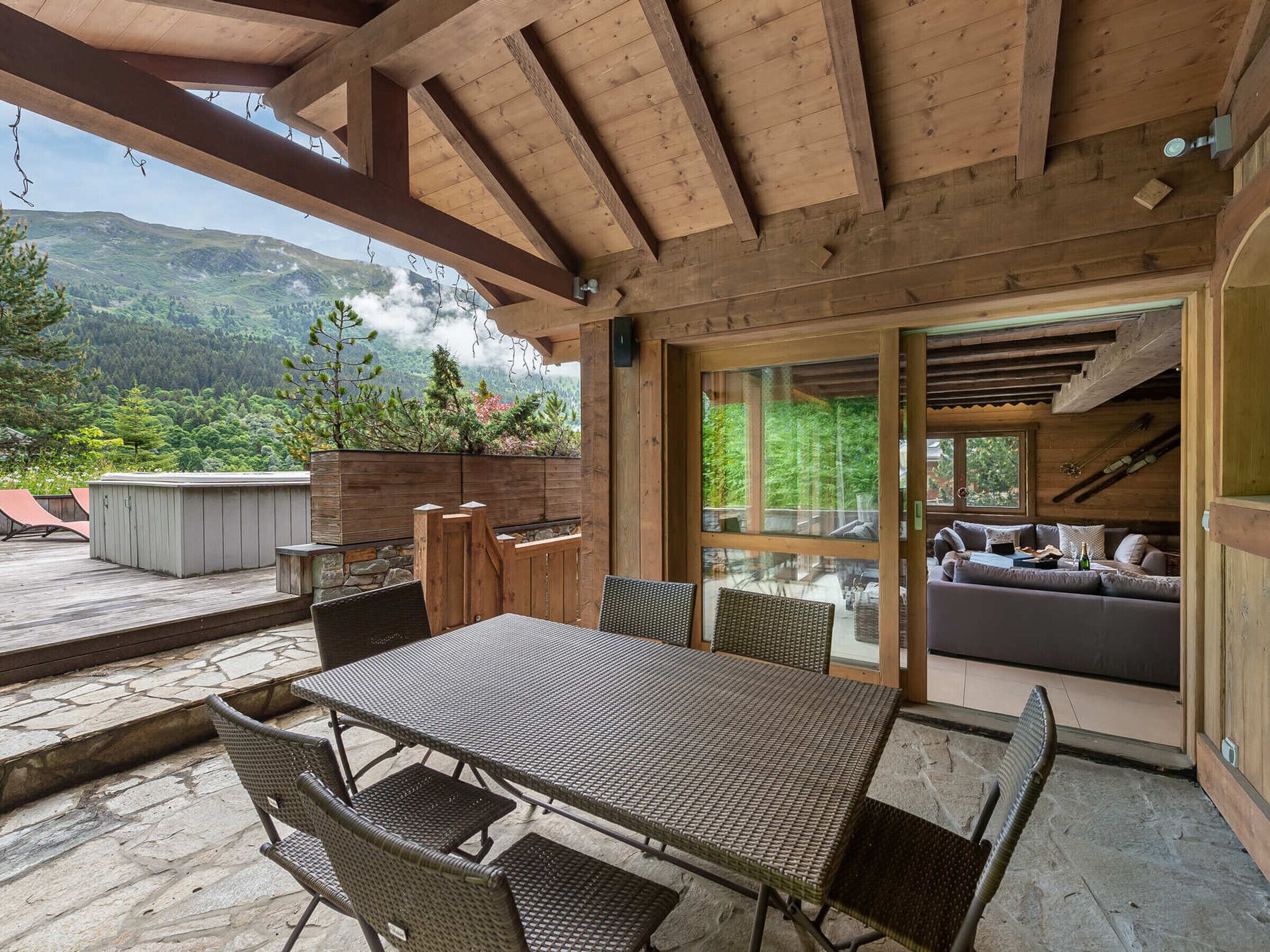 An outdoor patio with a wooden table and chairs, a hot tub, and a glass door leading to a living area with a sofa and a fireplace.