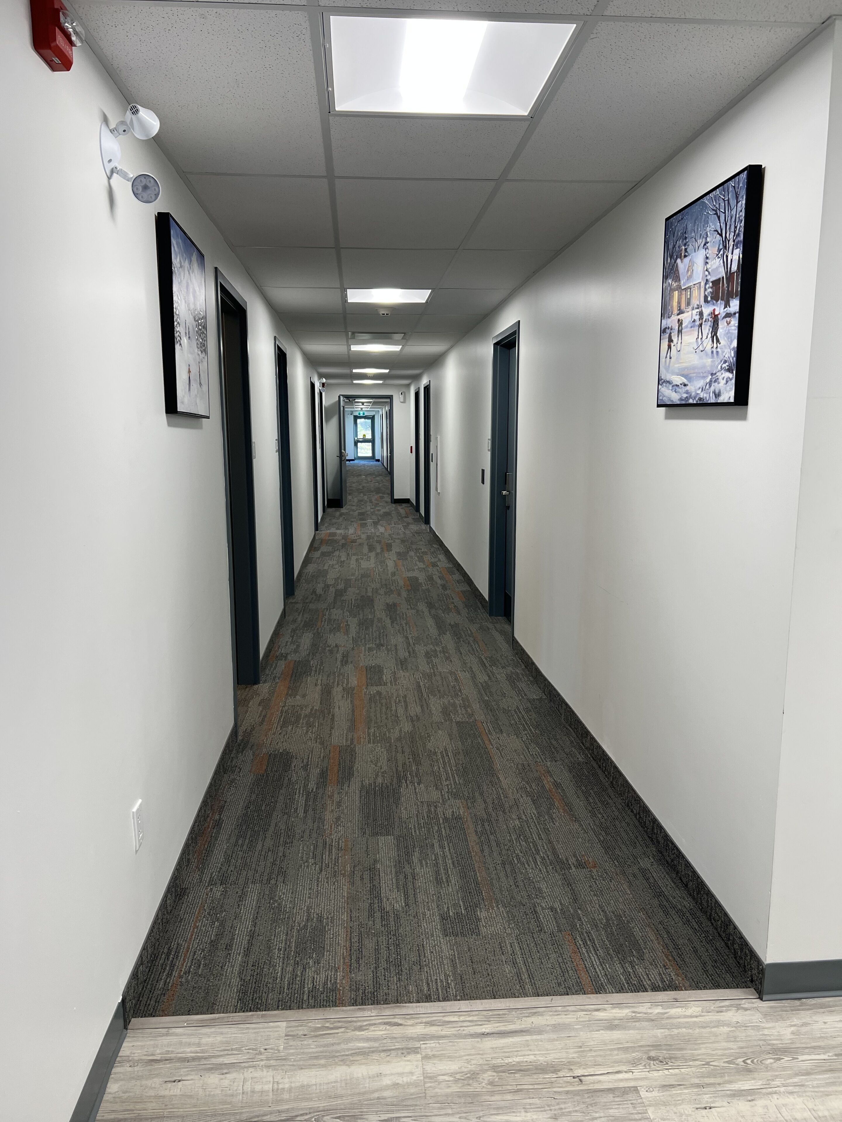 A long, narrow hallway with carpeted floors and white walls, featuring doors on both sides and a ceiling with fluorescent lights.