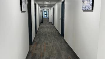 A long, narrow hallway with carpeted floors and white walls, featuring doors on both sides and a ceiling with fluorescent lights.