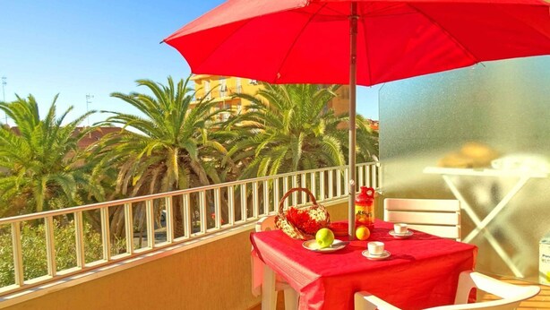 Un balcón con una sombrilla roja, sillas blancas, una mesa con mantel rojo y vistas a palmeras y edificios.