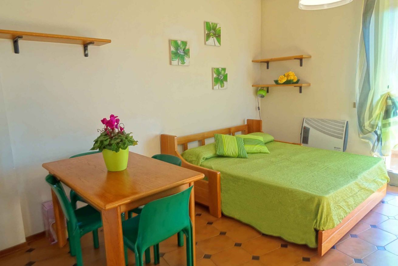 Ein kleiner, hell erleuchteter Raum mit einem hölzernen Bett, grüner Bettwäsche, einem Holztisch mit zwei Stühlen und einer Topfpflanze.