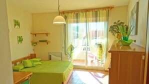 Una habitación con cama, escritorio, silla, radiador, ventana con cortinas y planta en el alféizar.