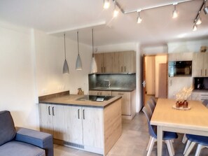 A modern kitchen with wooden cabinets, a central island, pendant lighting, and a dining area with blue chairs.