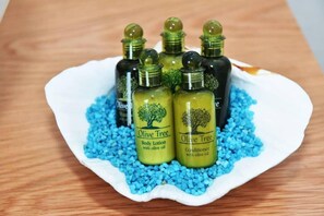 A white shell-shaped dish containing four bottles of Olive Tree body lotion and conditioner, each with a green label, surrounded by blue beads. - Christianna Luxury Apartment 2 (Apokoronas)