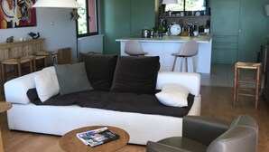 En moderne stue med sofa, sofabord og lænestole. Et køkken med grønne skabe og et barområde ses i baggrunden.