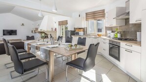 Et moderne køkken med hvide skabe, et spiseområde med et træbord og sorte stole, en indbygget ovn samt et stort vindue med persienner.