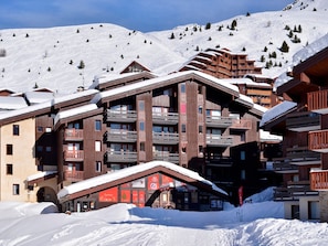Um hotel de vários andares com varandas de madeira e telhados cobertos de neve, com uma paisagem montanhosa nevada ao fundo.