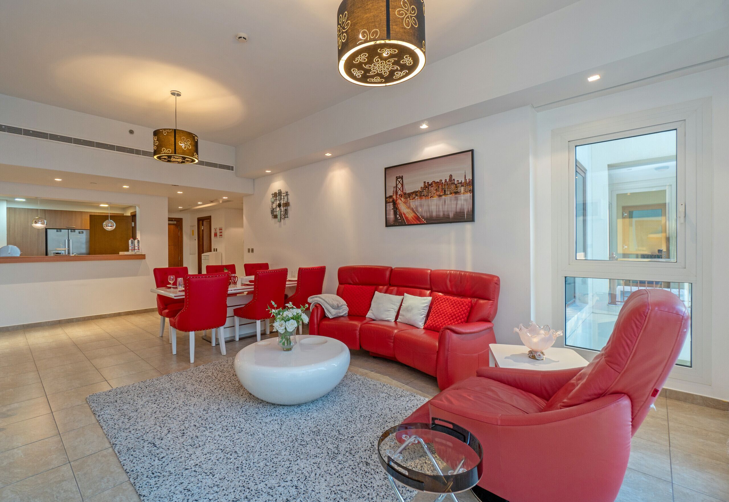 A modern living room with a red sofa, white coffee table, and red chairs. There is a painting on the wall and a large window providing natural light.
