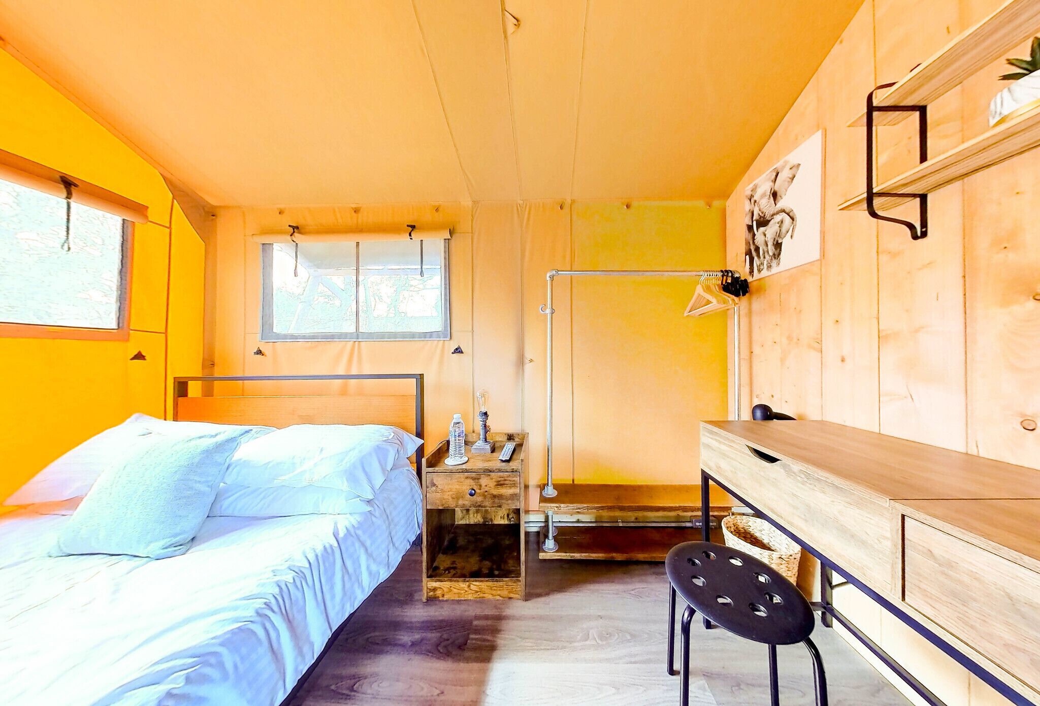 A compact room with a bed, a desk, and a chair. There is a window providing natural light, and a wall-mounted shelf with a lamp.