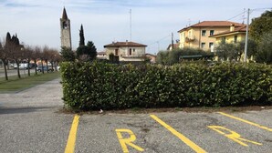 Ein Parkplatz mit zwei markierten Stellplätzen, einer Hecke und einem Gebäude mit einem Uhrturm im Hintergrund.