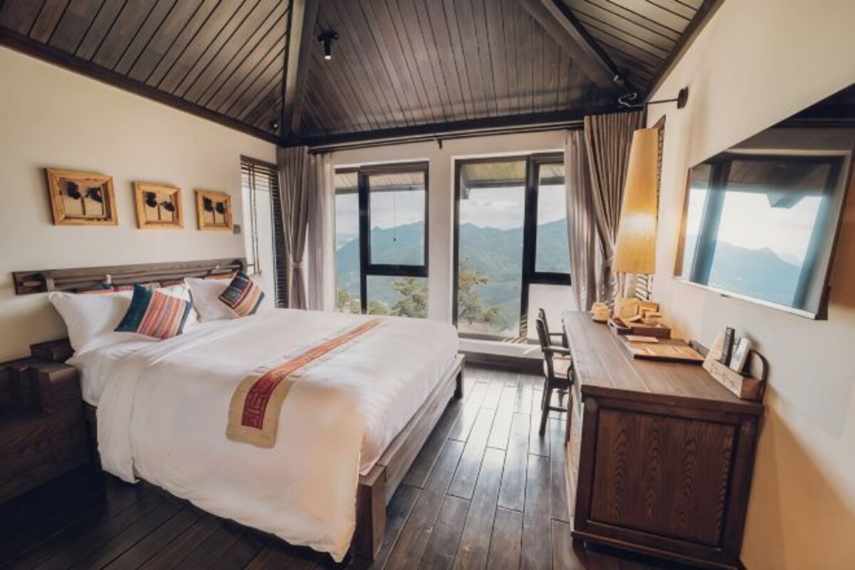 Une chambre à coucher avec un lit, des meubles en bois, un bureau et de grandes fenêtres donnant sur la montagne.