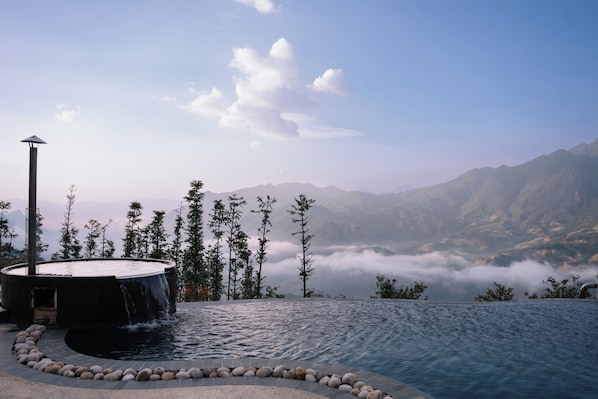 A infinity pool with a circular edge, surrounded by rocks and trees, overlooking a mountainous landscape with a waterfall.